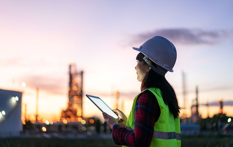 Asian female engineer in personal protective equipment using a digital tablet outside a refinery plant at sunset 