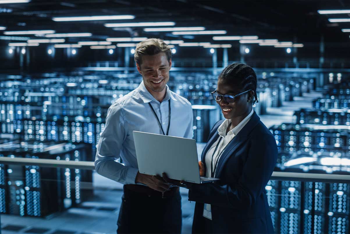 IT specialist and female IT specialist in the data centre, smiling and looking at a laptop