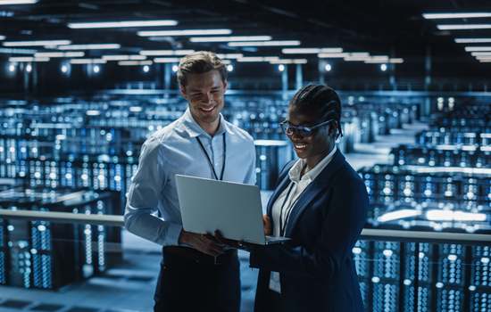 Two professionals standing in a server room, reviewing information on a laptop.
