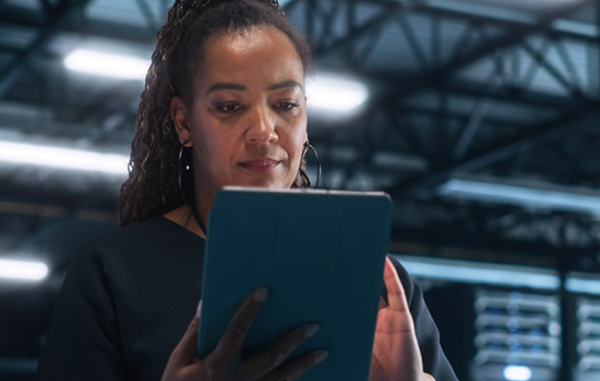Black woman holding and using a tablet in a large industrial or warehouse setting with metal beams and equipment in the background