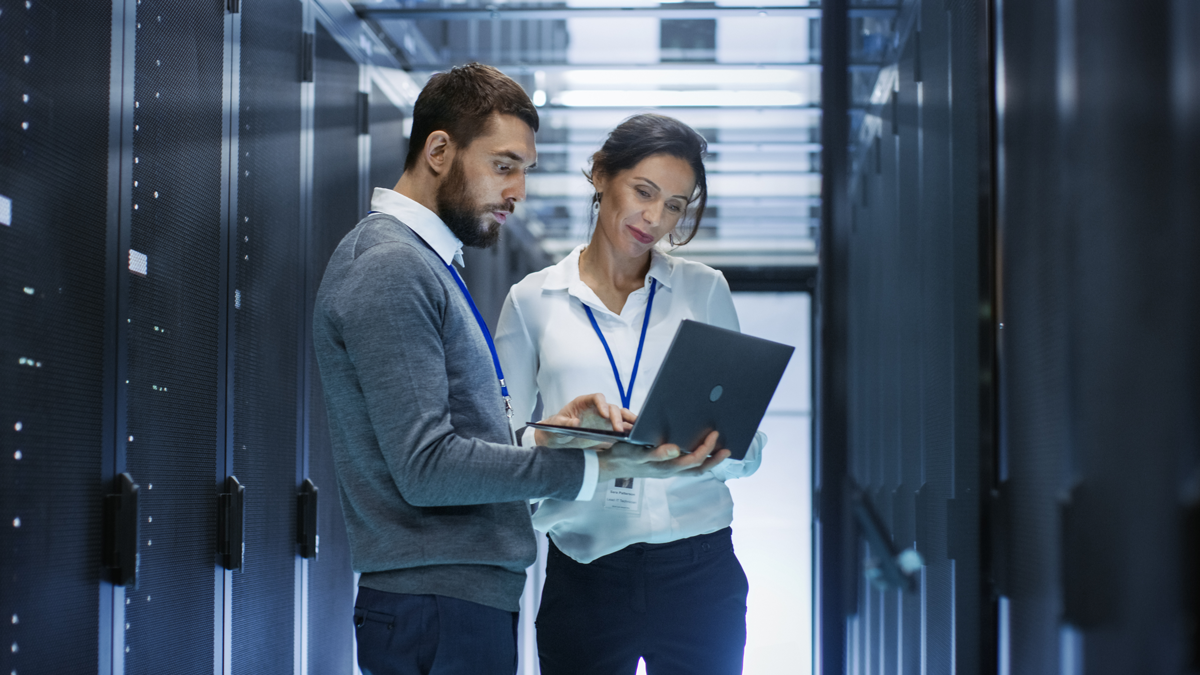 Male IT specialist holds laptop and discusses with female coworker in a data center whilst looking at IT racks