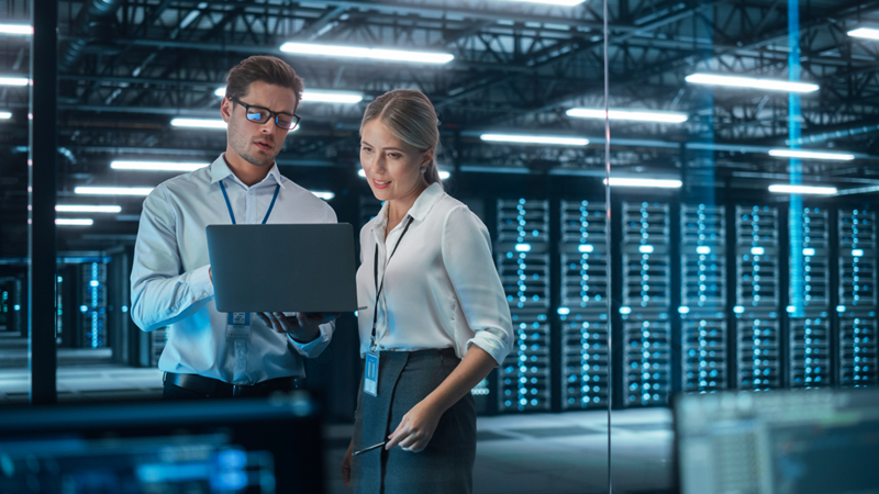 Two professionals standing in a server room, reviewing information on a laptop.