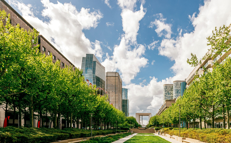 Straight parkway that blends urban buildings, lush green lawns, and blue skies