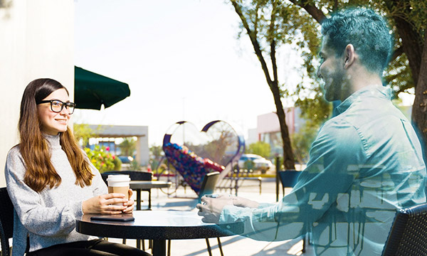 Woman drinking a coffee with a hologram of a man, depicting a borderless world created by physical and digital convergence.