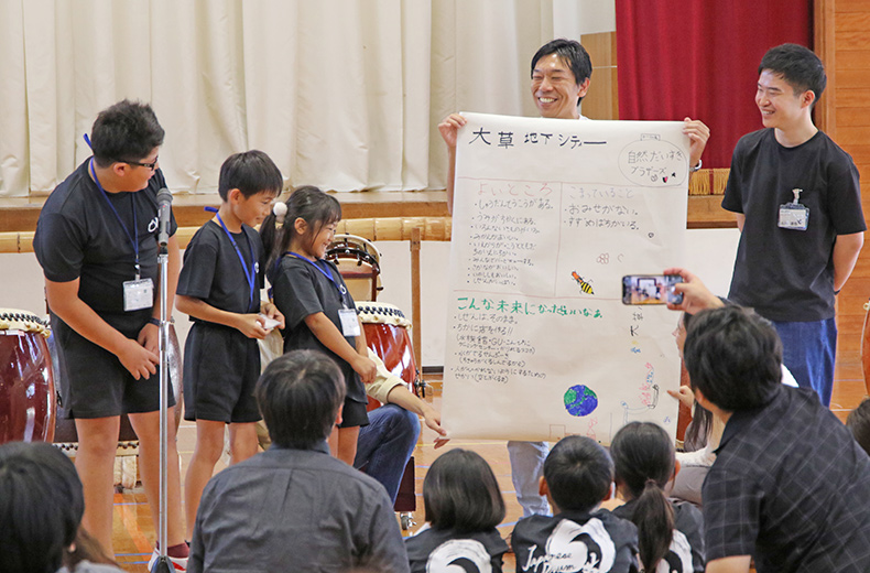 体育館にて、地域活性化のアイデアを描いたポスターを元に発表する小学生の様子