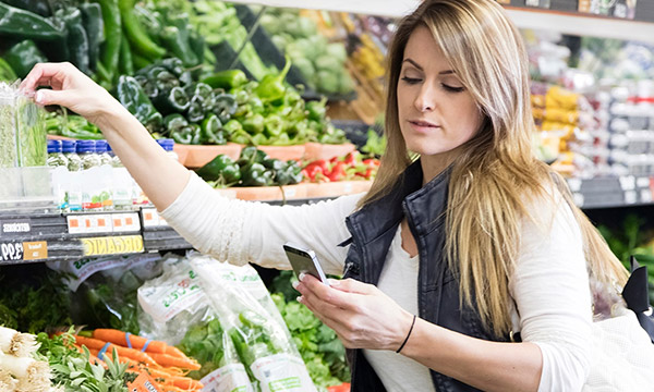 野菜売り場でスマホを見ている女性の画像。