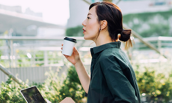 熱い飲み物を持った女性が座って、外でノートパソコンを操作している画像。